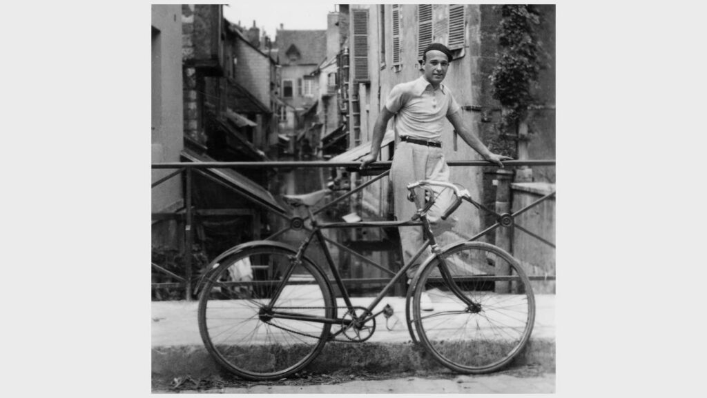 Mies polkupyörän kanssa pariisilaisen katumaiseman edessä. Kuva mustavalkoinen.