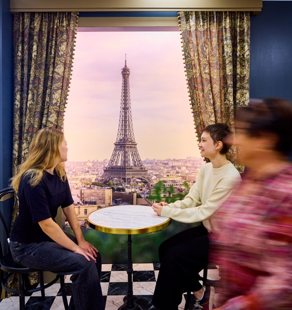 Kaksi nuorta aikuista istuu ranskalaista kahvilaa kuvaavassa lavasteessa kahvilan pöydässä, taustalla iso valokuva Pariisista.