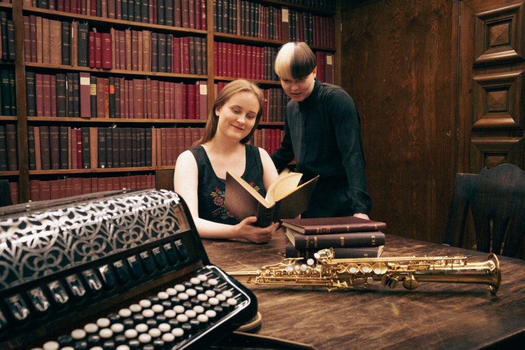 Kaksi muusikkoa kumartunut tutkimaan kirjaa arkistossa. Pöydällä harmonikka ja saksofoni.