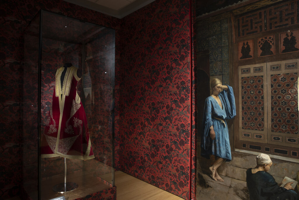 Tunnelmallisessa viininpunaisella tapetoidussa näyttelyhuoneessa oikealla näkyy orientalistinen Grommen maalaus, vasemmalla kultakirjailtu albanialainen viitta vitriinissä.