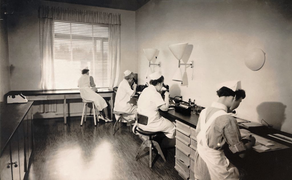 Naisia työskentelee seinää kiertävän työpöydän ympärillä työhönsä kumartuneena sairaalan laboratoriossa mustavalkoisessa kuvassa.