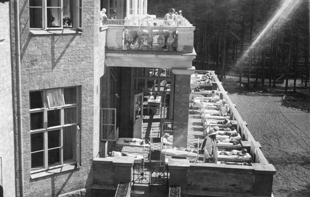 Parantolan julkisivu kuvattuna sivusta. Parvekeella lapsia makaa sängyssä ja hoitajia valkoisissa asuissaan.